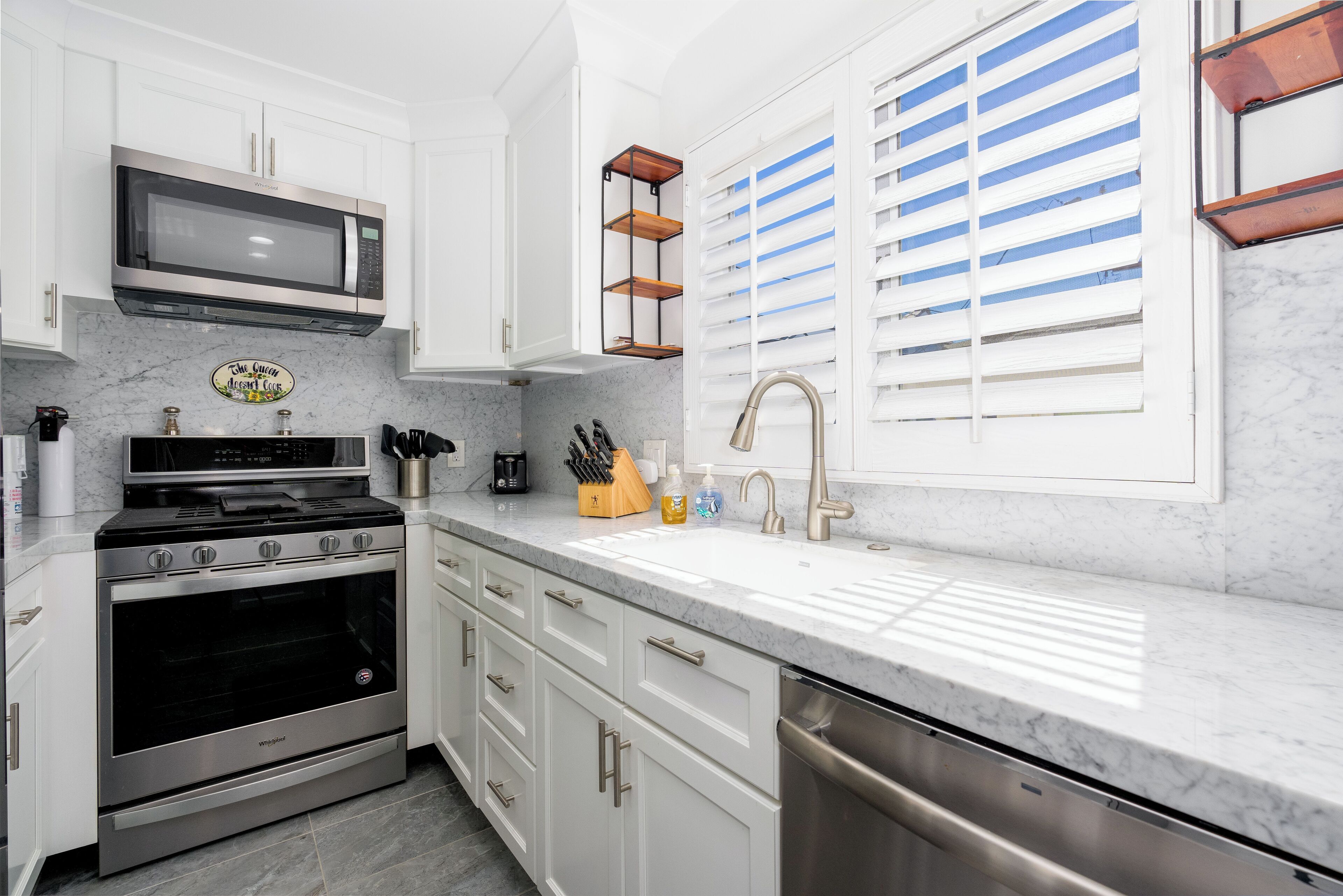 A modern kitchen with white cabinets, stainless steel appliances, a marble countertop, and a window with blinds.
