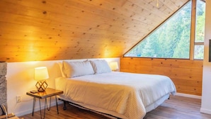 A bedroom with a wooden ceiling, a large window, a bed with striped bedding, and a nightstand with a lamp.