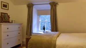Ein Schlafzimmer mit einem Bett, einem Schminktisch, einem Fenster mit Vorhängen und einem gerahmten Bild auf dem Schminktisch.