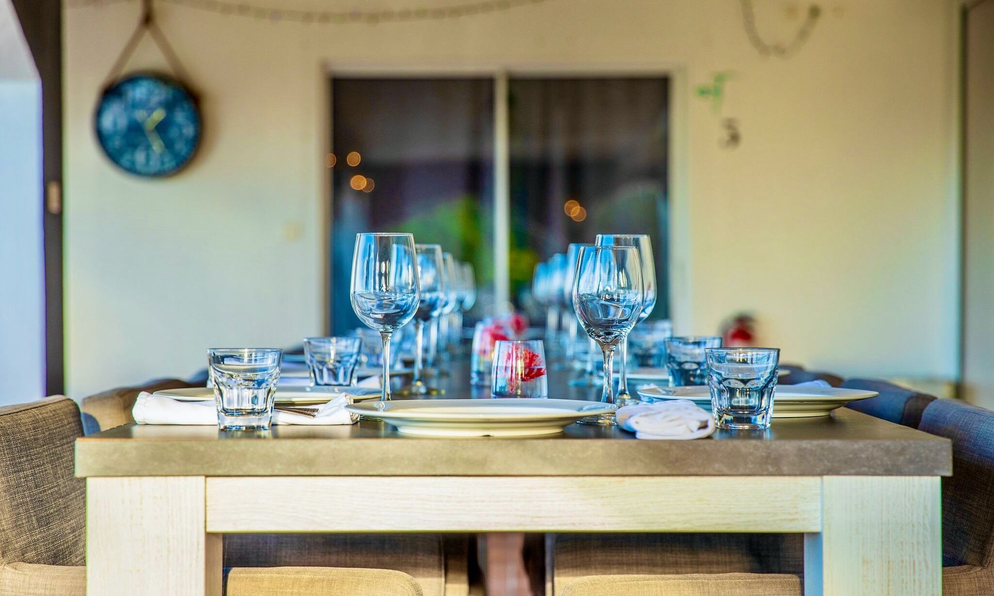 A dining table set with multiple wine glasses and plates, a clock on the wall, and a window in the background.