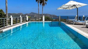 A clear blue pool with a wooden deck, white railings, and a view of the ocean and mountains in the background.