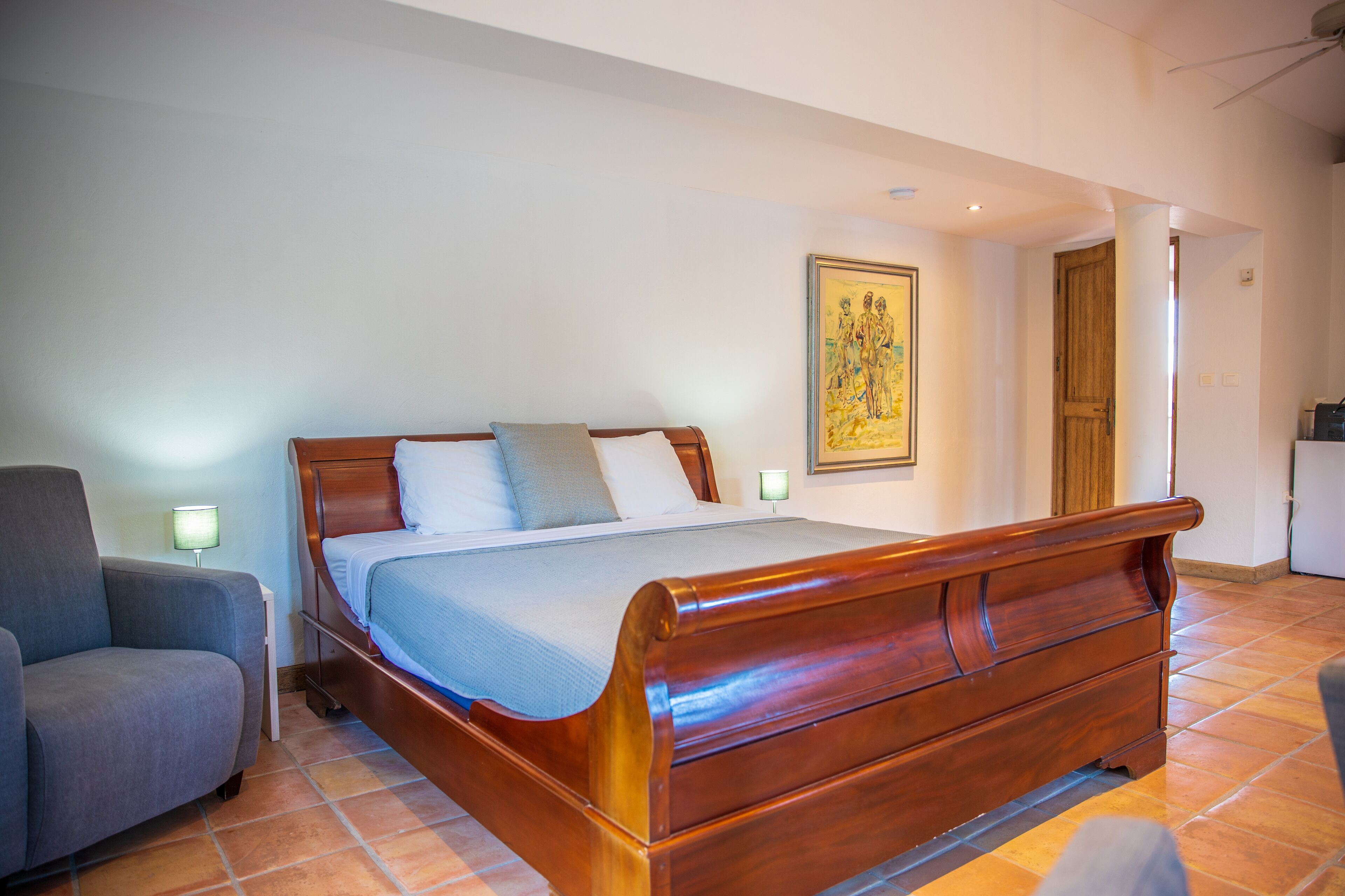 A bedroom with a wooden bed, a grey armchair, and a nightstand with a lamp.