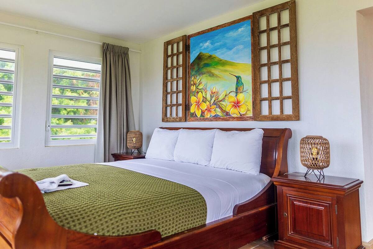 A neatly made bed with a green patterned bedspread, a wooden headboard, and white pillows. There is a painting of a tropical scene above the bed.
