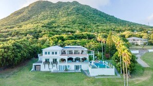 A large, two-story house with a swimming pool, surrounded by lush greenery and a mountain in the background.
