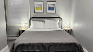 A bedroom with a bed, two bedside tables, a window with blinds, and two framed pictures on the wall. - Cozy & Comfy 1 Bedroom Apartment Downtown Sault, ON (Sault Ste. Marie)