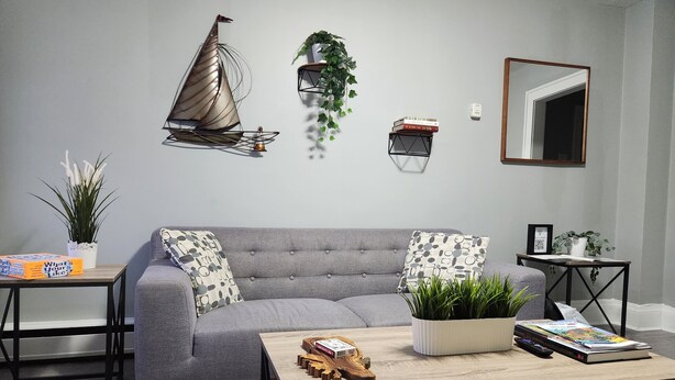 A living room with a grey sofa, a coffee table, a wall-mounted sailboat decoration, a small plant, and a bookshelf. - Cozy & Comfy 1 Bedroom Apartment Downtown Sault, ON (Sault Ste. Marie)