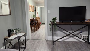 A room with a TV on a wooden stand, a small table with a plant, and a doorway leading to a dining area with a table and chairs. - Cozy & Comfy 1 Bedroom Apartment Downtown Sault, ON (Sault Ste. Marie)