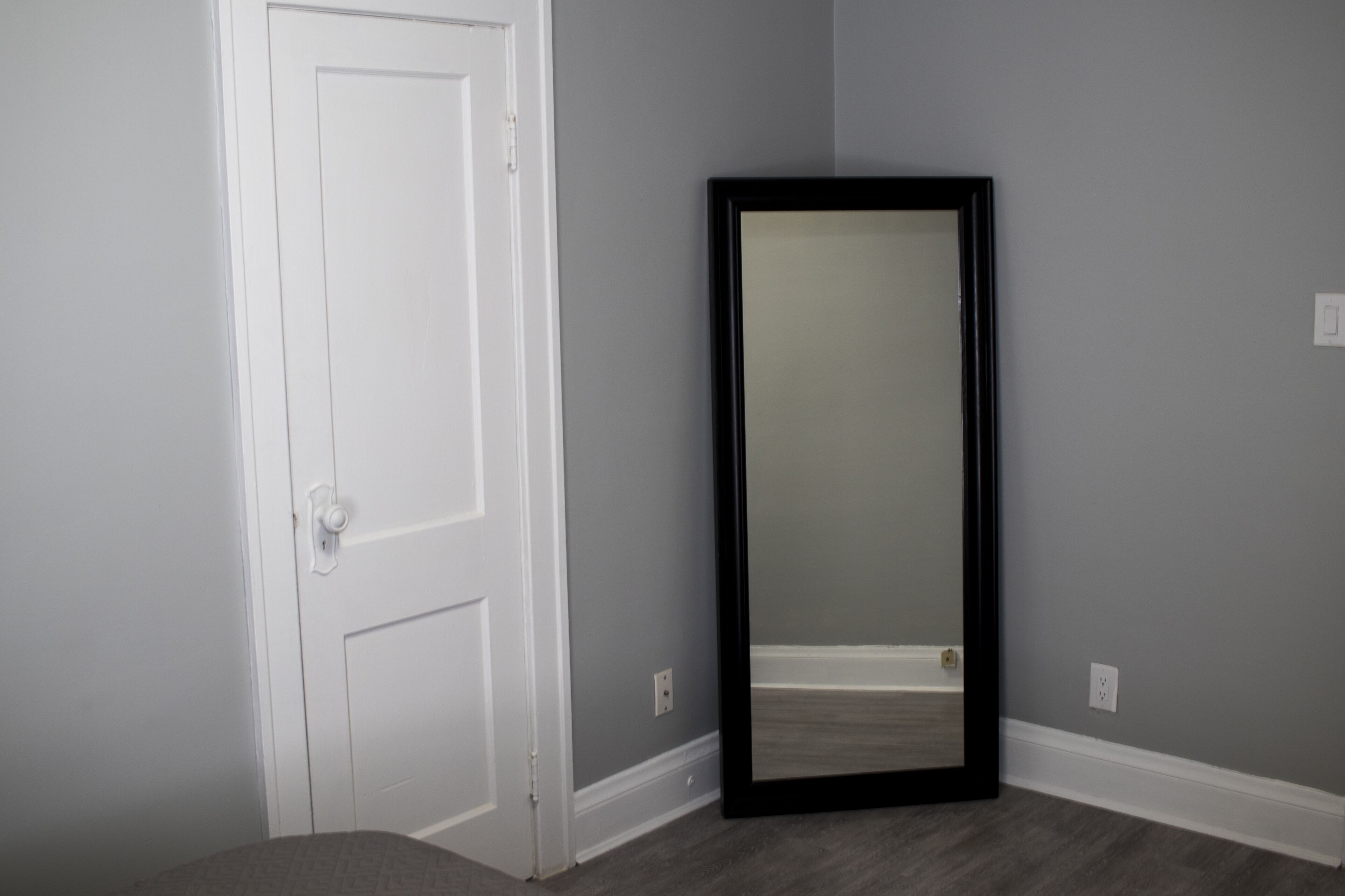 Una habitación con una puerta blanca cerrada, un espejo de marco negro y paredes grises.