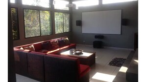 A living room with a red sofa, a coffee table, a projector screen, and large windows with a view of trees. - Tropical Paradise. Little Corn Island (RAAS)
