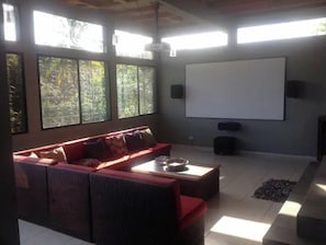 A living room with a red sofa, a coffee table, a projector screen, and large windows with a view of trees. - Tropical Paradise. Little Corn Island (RAAS)
