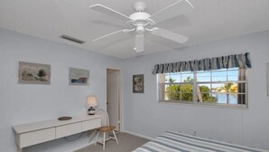 Un dormitorio con cama, escritorio, silla, ventilador de techo y ventana con vista a palmeras.