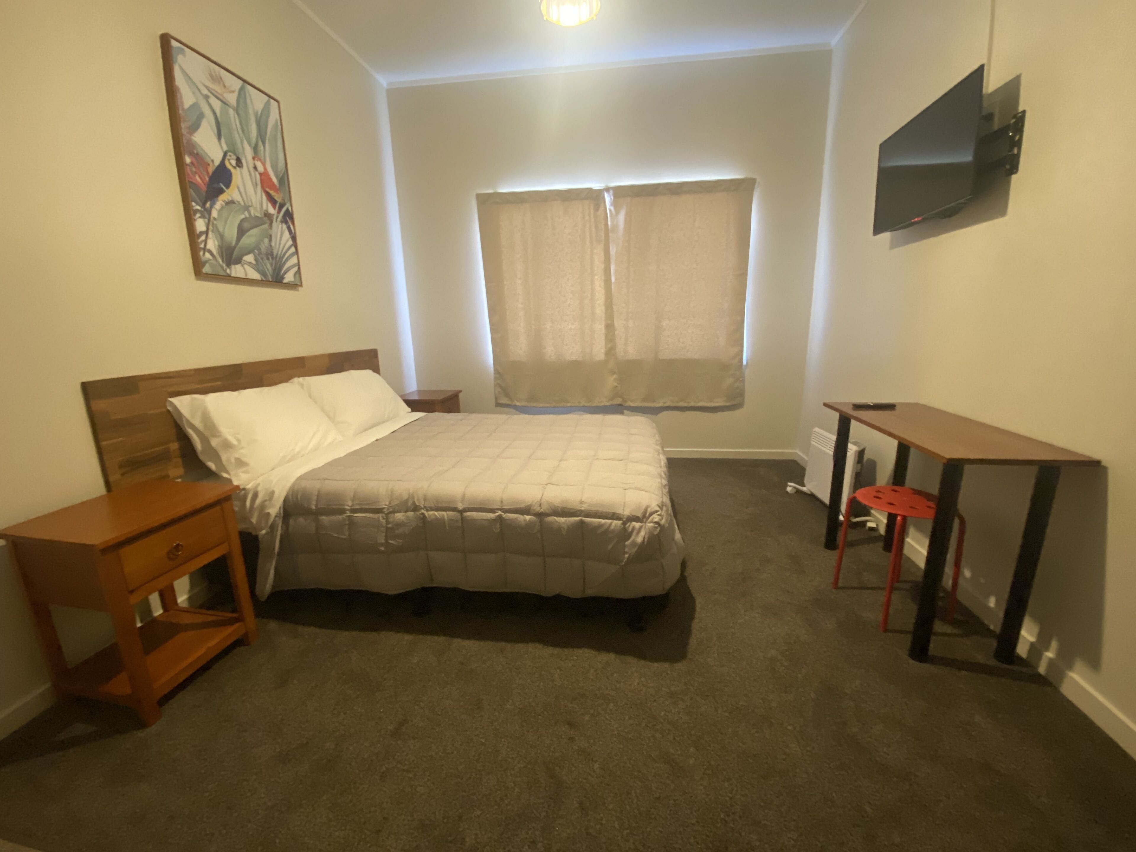 Quarto com cama, escrivaninha, cadeira, televisão, janela com cortinas e um quadro na parede.
