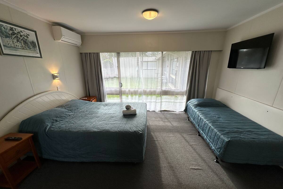 Une chambre d’hôtel avec deux lits, une petite table de chevet, un téléviseur fixé au mur et une fenêtre avec des rideaux.