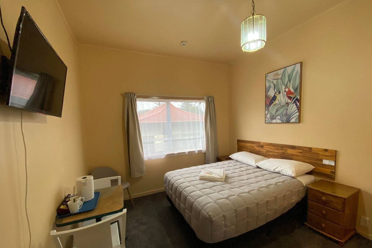 Une chambre à coucher avec un lit, un bureau et une chaise, un téléviseur fixé au mur, une fenêtre avec des rideaux et un tableau accroché au mur.