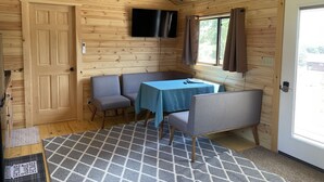 A cozy cabin room with a wooden interior, a table with chairs, a TV mounted on the wall, and a door leading outside. - Cooperstown Double Play Hilltop Cabin B (Cooperstown)