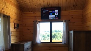 A wooden cabin room with a television mounted above a window, a wooden dresser, and a small black trash bin. - Cooperstown Double Play Hilltop Cabin B (Cooperstown)