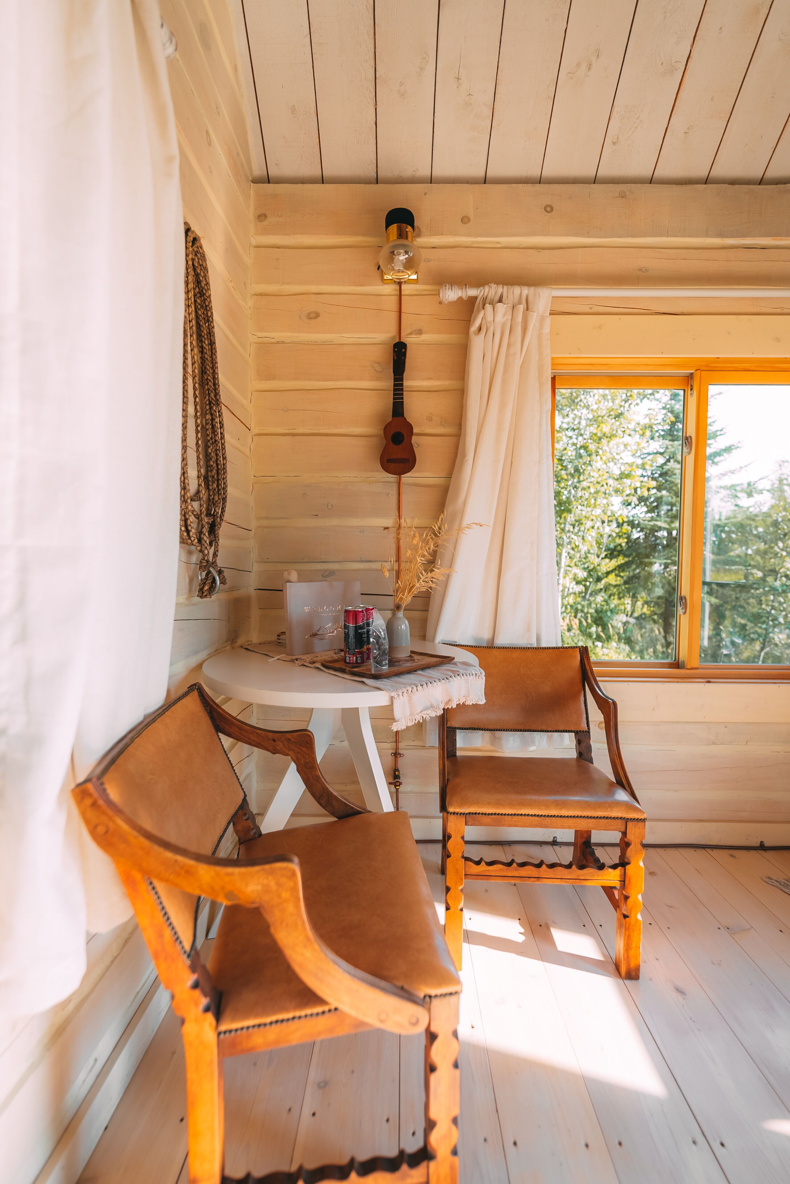 En stuga med träpanel på insidan, med två trästolar, ett litet runt bord, en gitarr som hänger på väggen och ett fönster med utsikt över träd.