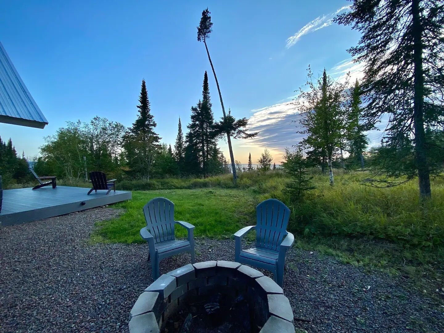 A deck with two Adirondack chairs facing a fire pit, surrounded by a natural landscape with trees and a clear sky.