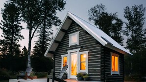 Une cabane en rondins avec un porche, deux chaises et un petit foyer extérieur.