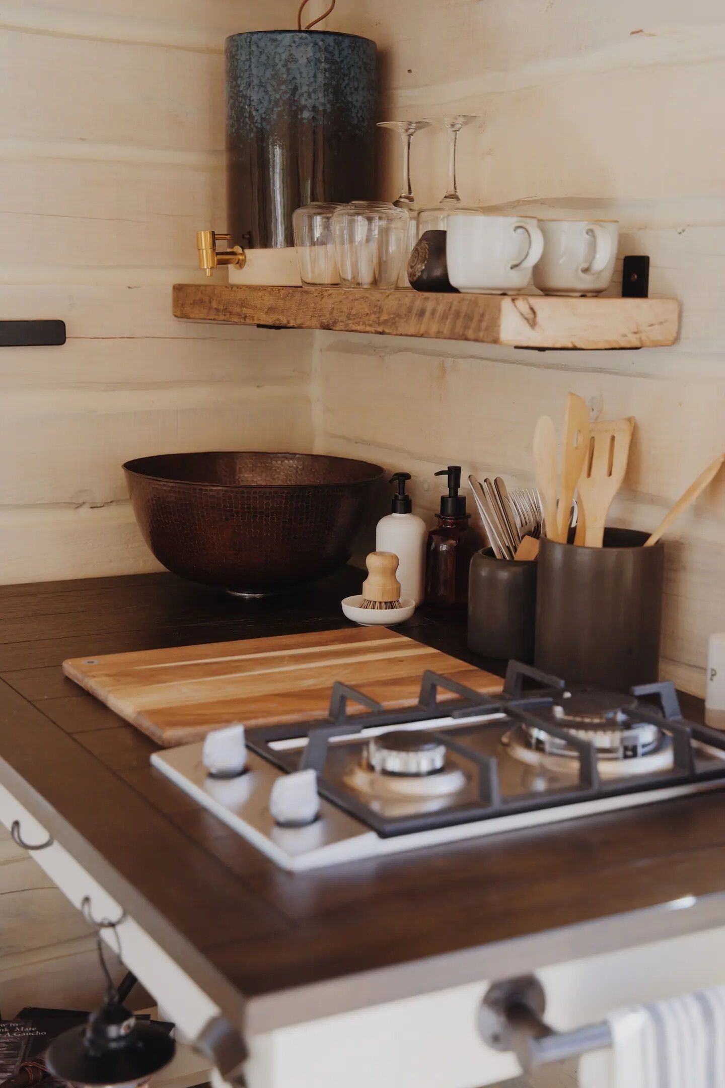 A kitchen with a gas stove, wooden cutting board, metal bowl, and utensils on a shelf.