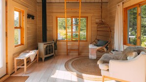 L’intérieur d’un chalet accueillant, avec un plafond en bois, une cheminée, un fauteuil suspendu, un canapé et une grande fenêtre donnant sur le paysage extérieur.