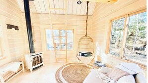 Un intérieur de chalet chaleureux avec une grande fenêtre, un fauteuil suspendu, une cheminée et un canapé blanc.