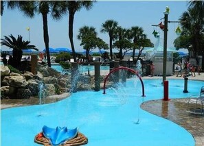 Relax by the clear blue pool surrounded by palm trees and a rocky landscape, where a large fountain and water slide create a soothing atmosphere.