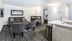Una sala moderna con un sofá gris, dos sillones, una mesa de comedor con taburetes altos y una cocina con encimera blanca y fregadero.