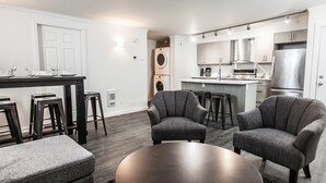 Una cocina moderna con electrodomésticos de acero inoxidable, una zona de comedor con taburetes altos y un rincón de estar con dos sillones y una mesa redonda.