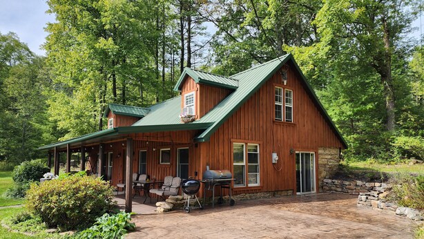 A wooden cabin with a covered patio, stone foundation, and a green roof, surrounded by lush greenery. - Charming re-imagined bank barn cabin. (Millmont)