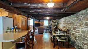 A rustic kitchen with wooden beams, a stone wall, and a dining area with metal chairs. - Charming re-imagined bank barn cabin. (Millmont)