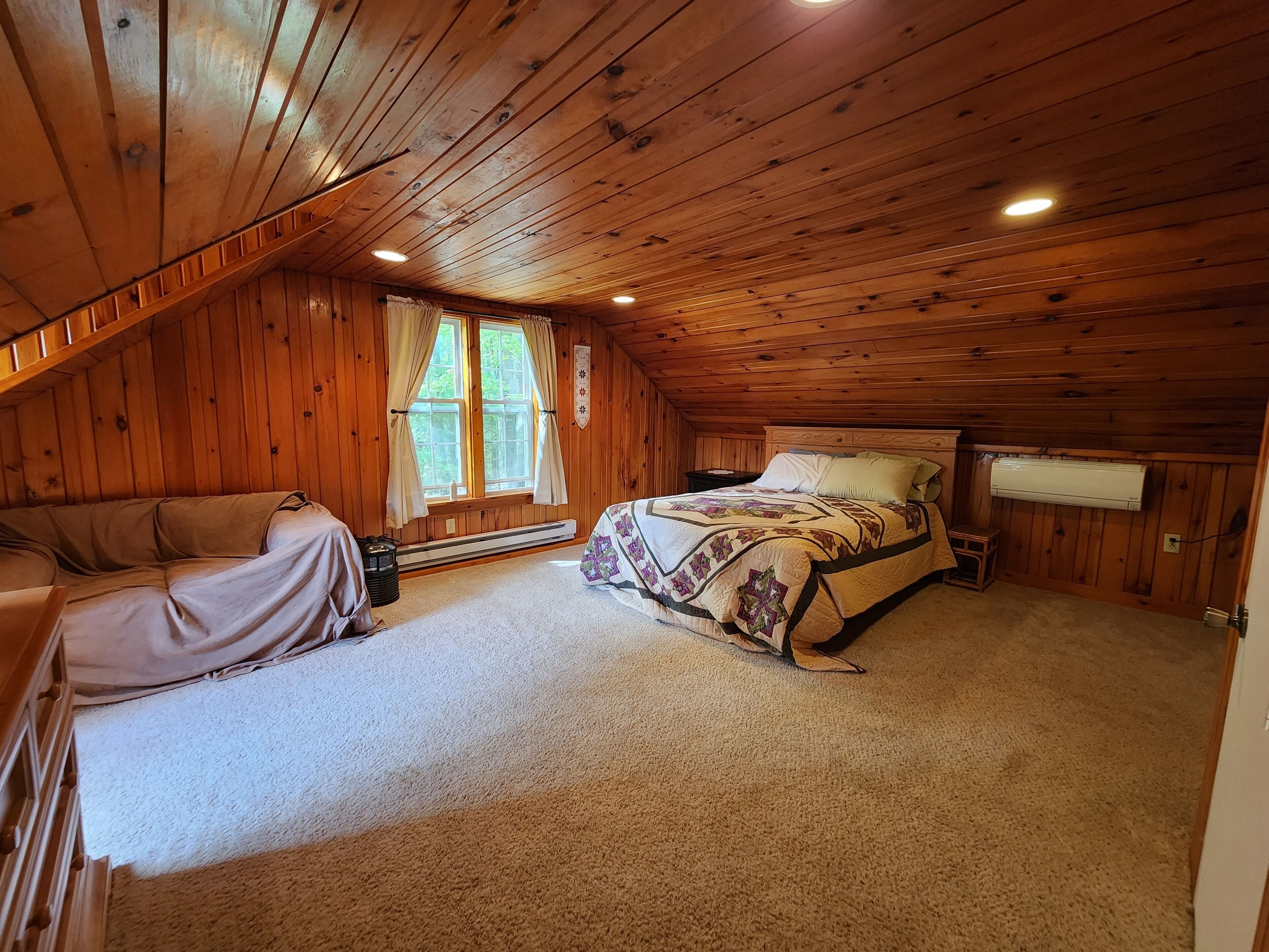A cozy attic bedroom with a wooden ceiling, a large bed with a patterned quilt, a nightstand, and a window with curtains.