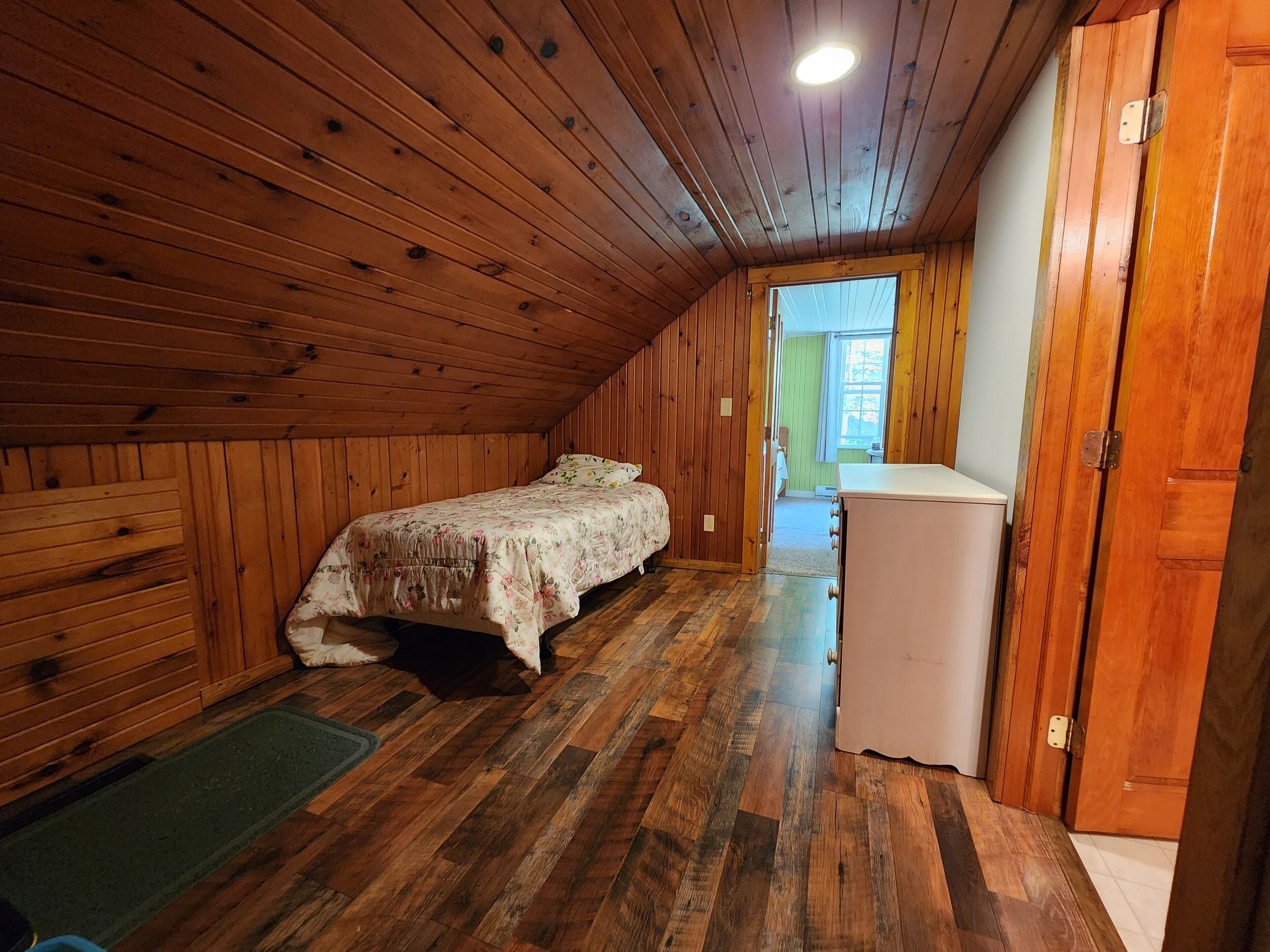 A wooden-paneled attic bedroom with a bed, a small white refrigerator, and a door leading to a hallway.