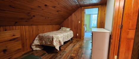 A wooden-paneled attic bedroom with a bed, a small white refrigerator, and a door leading to a hallway.