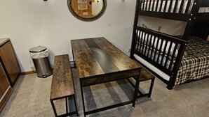 Una cama litera con colchón a cuadros, una mesa de madera con bancos, un cubo de basura y un espejo fijado a la pared.