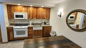 Cocina funcional con armarios de madera, microondas, estufa y lavavajillas. Se observa un espejo redondo en la pared y una mesa de madera oscura.