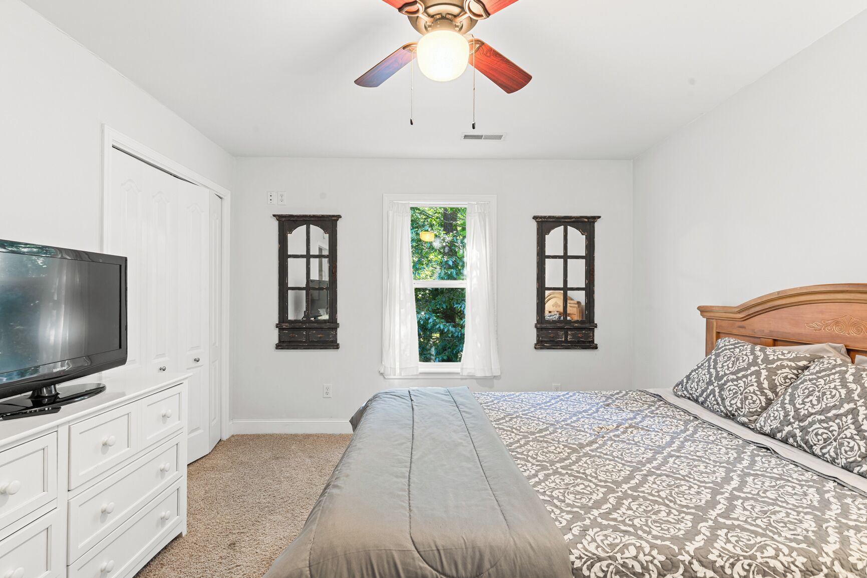 Une chambre à coucher avec un lit, une télévision, un ventilateur de plafond et deux fenêtres avec des rideaux.