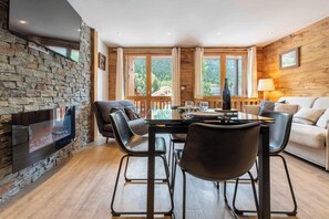 Ein gemütliches Wohnzimmer mit Kamin aus Stein, einem Esstisch mit Stühlen und Blick ins Freie durch große Fenster.