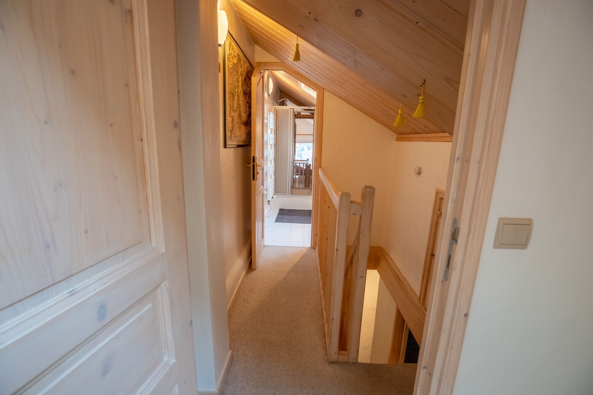 A narrow hallway with wooden walls and a sloped ceiling. A door on the left leads to a room with a large window. A small wooden shelf is visible on the right wall.