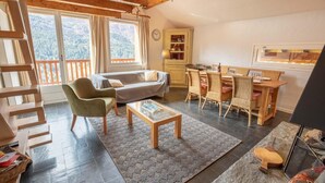 Ein gemütliches Wohnzimmer mit Sofa, Sessel und Esstisch. Eine Treppe ist links zu sehen, und ein Fenster bietet Blick auf Bäume.