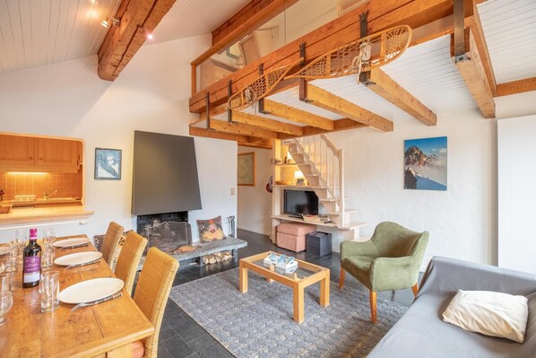 Amplia sala de estar con techo de madera, chimenea, mesa de comedor preparada para cuatro personas y un conjunto de asientos cómodo.