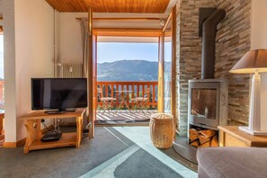 Una camera accogliente con TV, camino, balcone con sedie e vista sulle montagne.