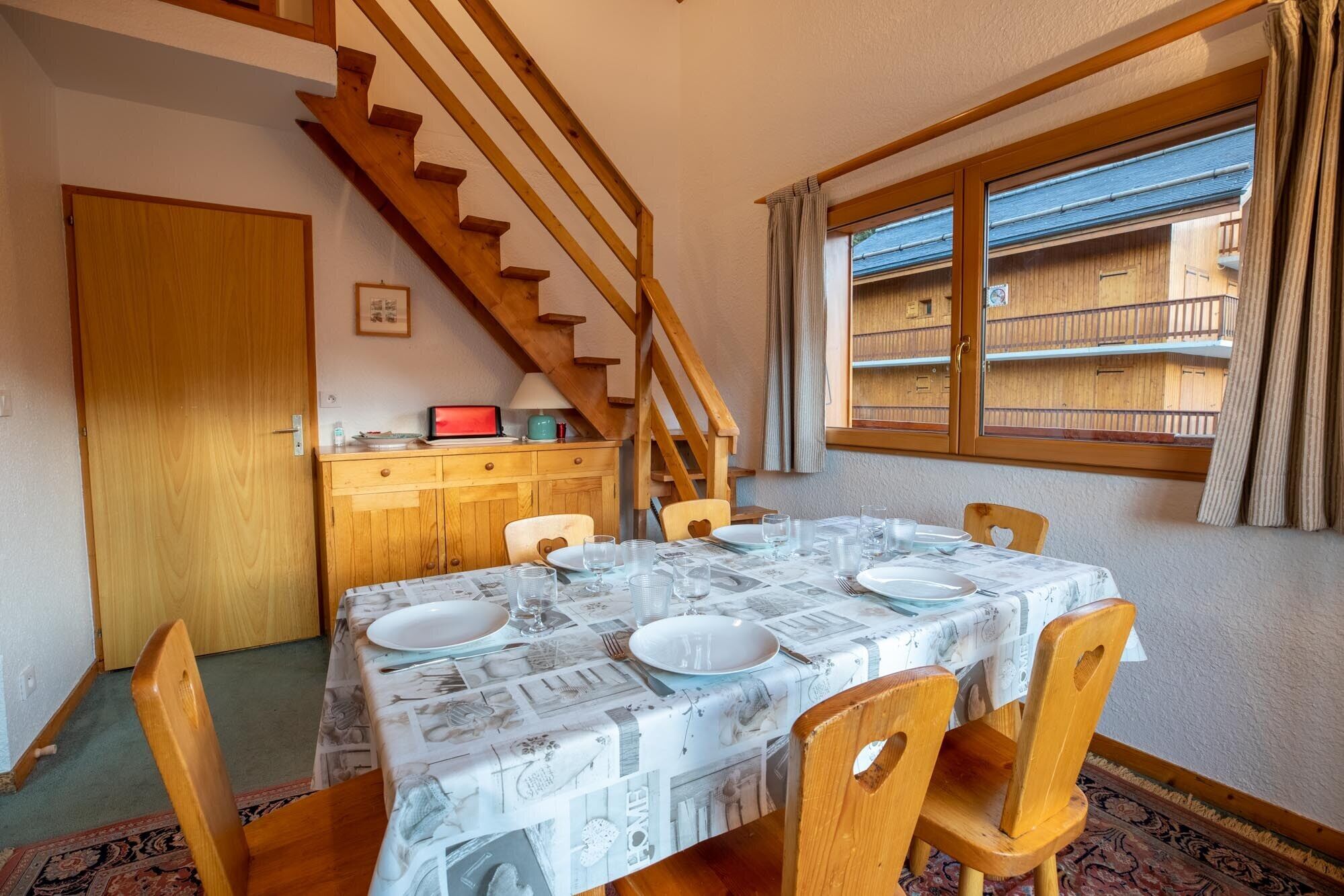 Zona pranzo con tavolo imbandito per quattro, sedie di legno, una scala e una finestra vista balcone.