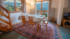 Een eetruimte met een tafel voor vier personen, houten stoelen, een kamerbreed tapijt met dessin en uitzicht op bergen door grote ramen.