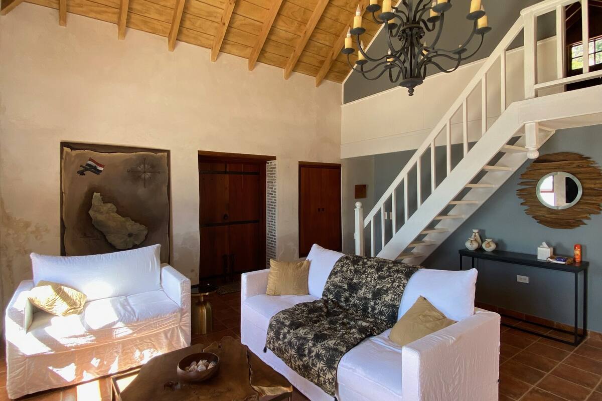 A spacious living room with a high wooden ceiling, a chandelier, and a staircase leading to an upper level.