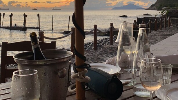 A table set for dining with a view of the sea at sunset, a metal pot, and several wine glasses.