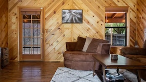 A cozy cabin interior with a wooden hipped roof, a fireplace, a sofa, and a large window. - Dog Friendly, Fully Gated, Private Cabin by Fool Hollow Lake (Show Low)