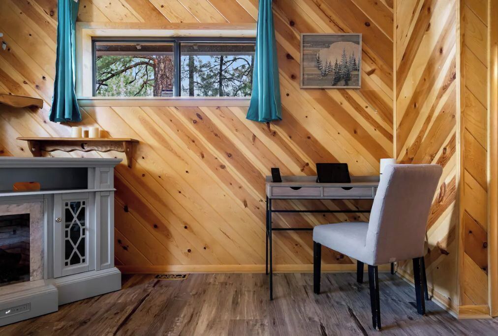 A cozy cabin room with a wooden interior, a desk with a chair, a fireplace, and a window with curtains.