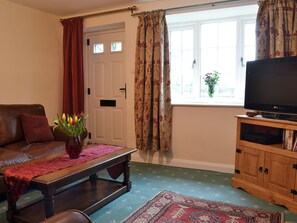 A living room with a brown leather sofa, a wooden coffee table with a vase of flowers, a television on a wooden stand, and a window with curtains. - Keldholme Cottage 1 - UK10992 (Keldholme, near Kirkbymoorside)
