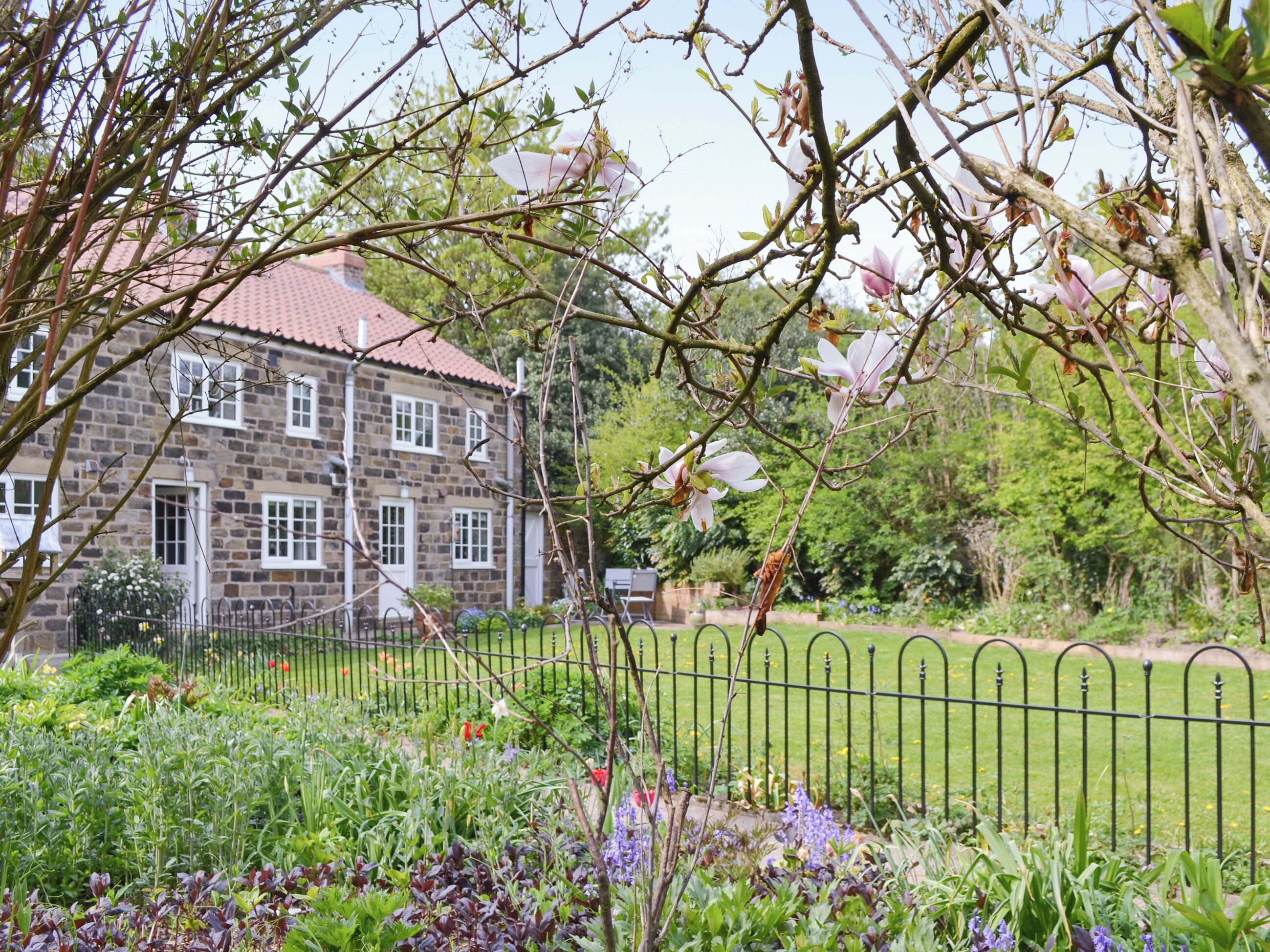 Een gebouw van steen met een rode dakbedekking, omgeven door een goed onderhouden tuin met diverse planten en een metalen hek.